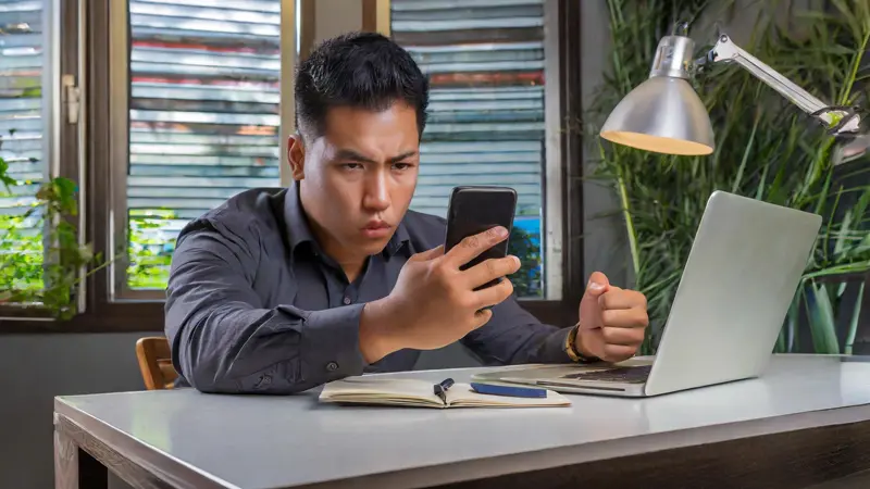 Stressed man at desk with laptop, intense gaze at phone with clenched fist – conveying emotions of anger and potential retaliation in a work-related claim.